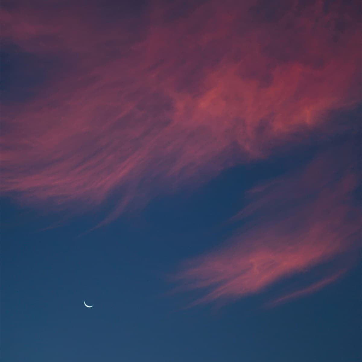Pink and purple clouds in a blue sky with a crescent moon.